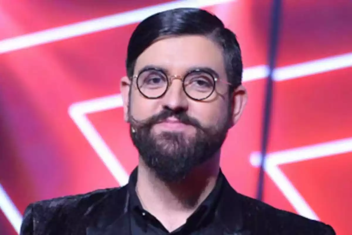 Manu Sánchez con barba y gafas redondas en un escenario con fondo rojo.