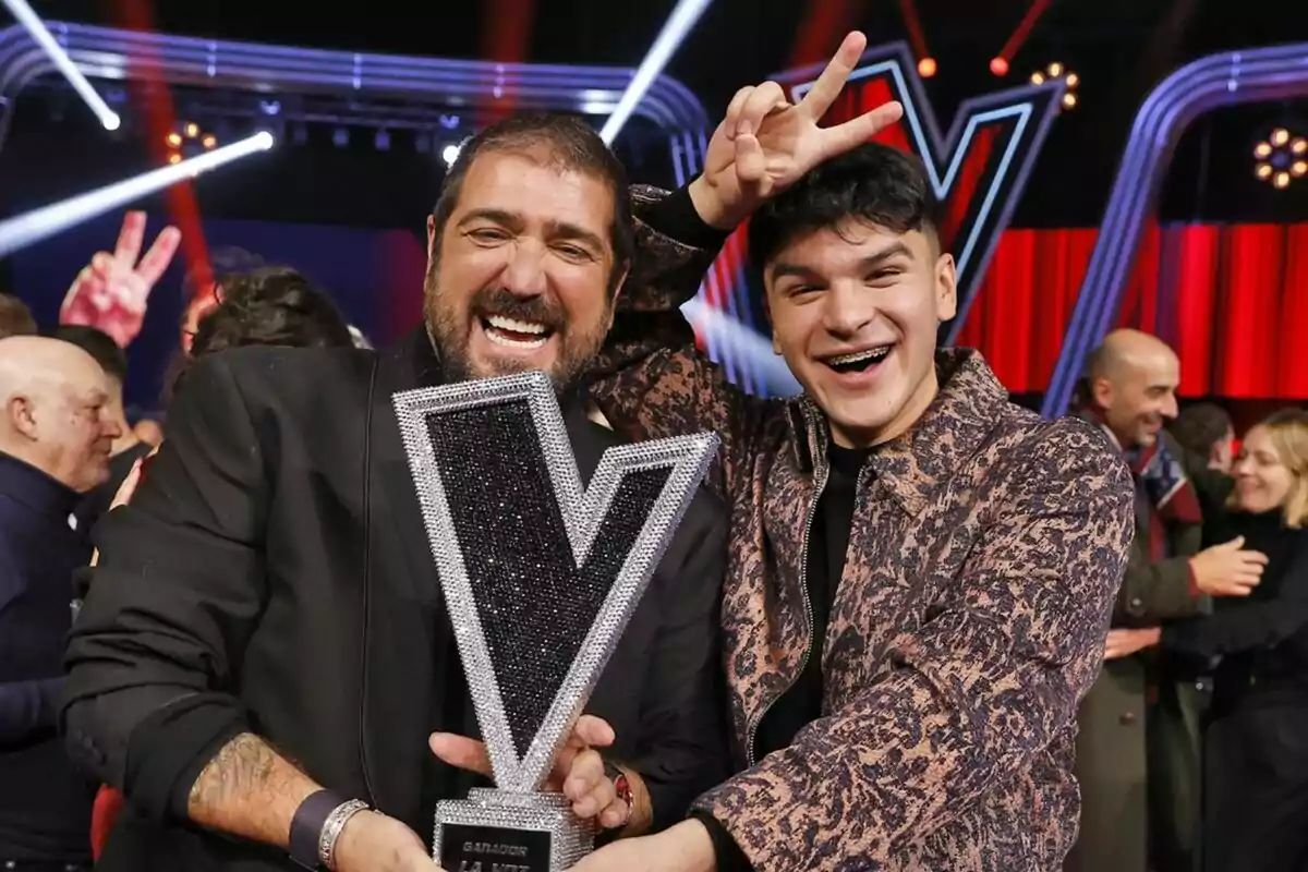 Antonio Orozco y Manuel Ayra sonrientes sostienen un trofeo de La Voz en un escenario iluminado.