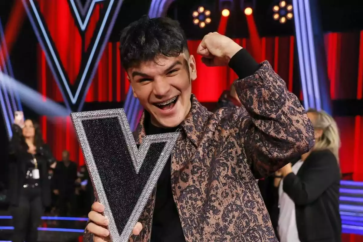 Manuel Ayra sonriente levanta un trofeo de La Voz en forma de V en un escenario iluminado con luces rojas y azules.