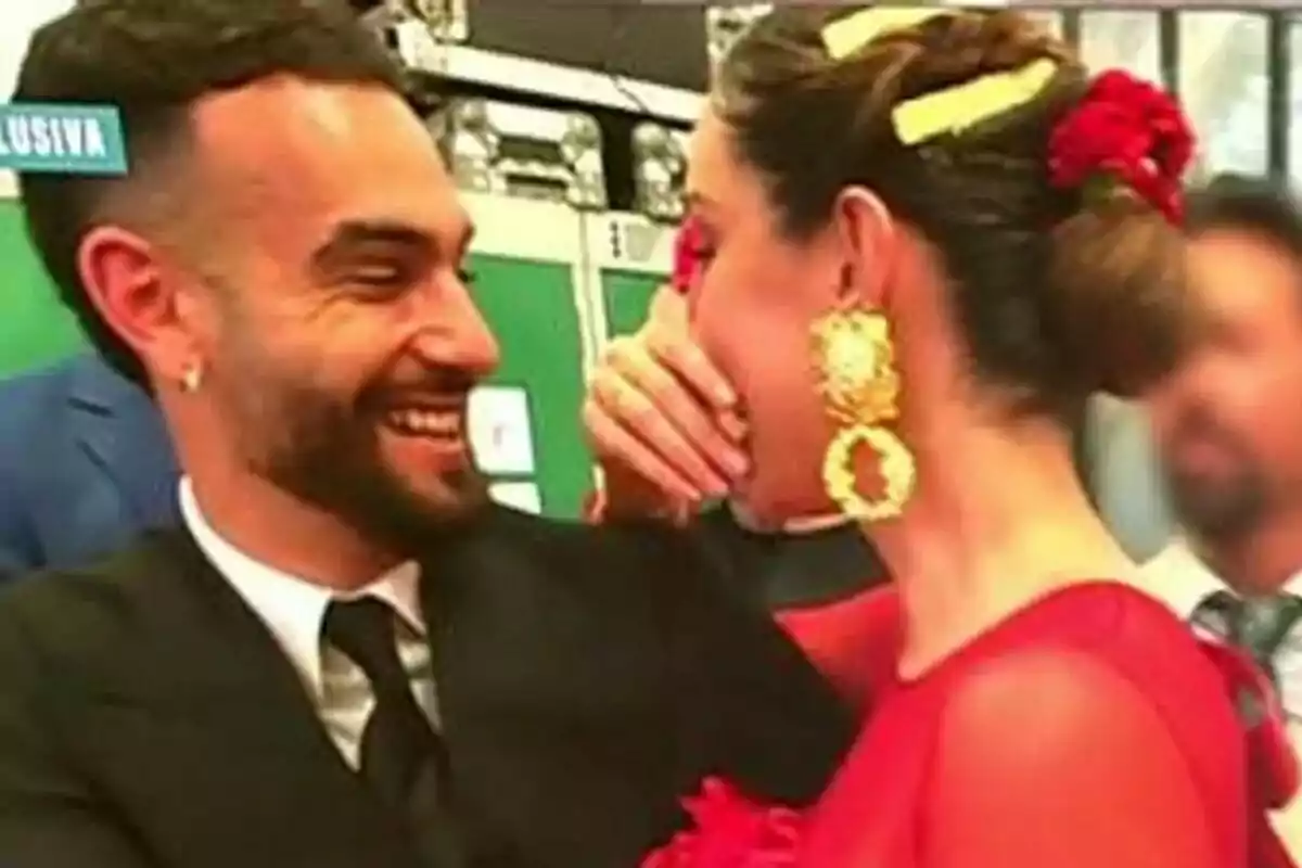 Manuel Cortés y Adara Molinero sonriente comparte un momento de alegría, con la mujer vistiendo un vestido rojo y pendientes dorados, mientras el hombre lleva un traje oscuro.
