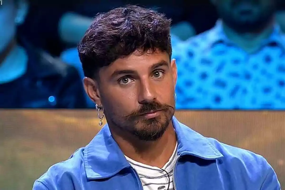 Manuel González con barba y bigote lleva una chaqueta azul y un pendiente en la oreja mientras está sentado en un entorno de estudio en el plató de Supervivientes con luces azules de fondo.