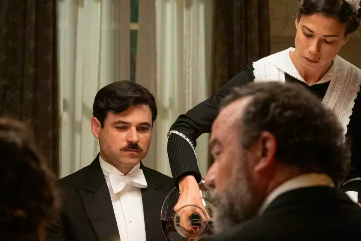 Una camarera sirve vino a Manuel con bigote que está sentado en una mesa elegantemente vestida, mientras otro hombre de barba está en primer plano en La Promesa.