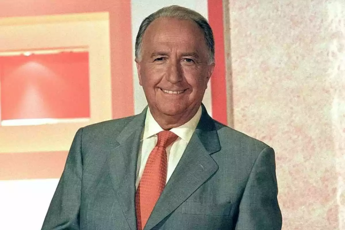 Manuel Torreiglesias mayor con traje gris y corbata naranja sonríe frente a un fondo con tonos rosados y blancos.
