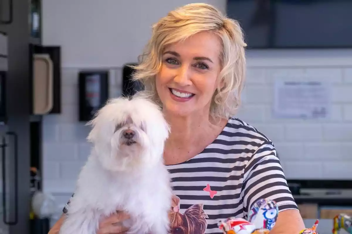 Mar Vega sonriente con cabello rubio corto sostiene a un perro pequeño y peludo en sus brazos, ambos están en un entorno interior en Patas Arriba.