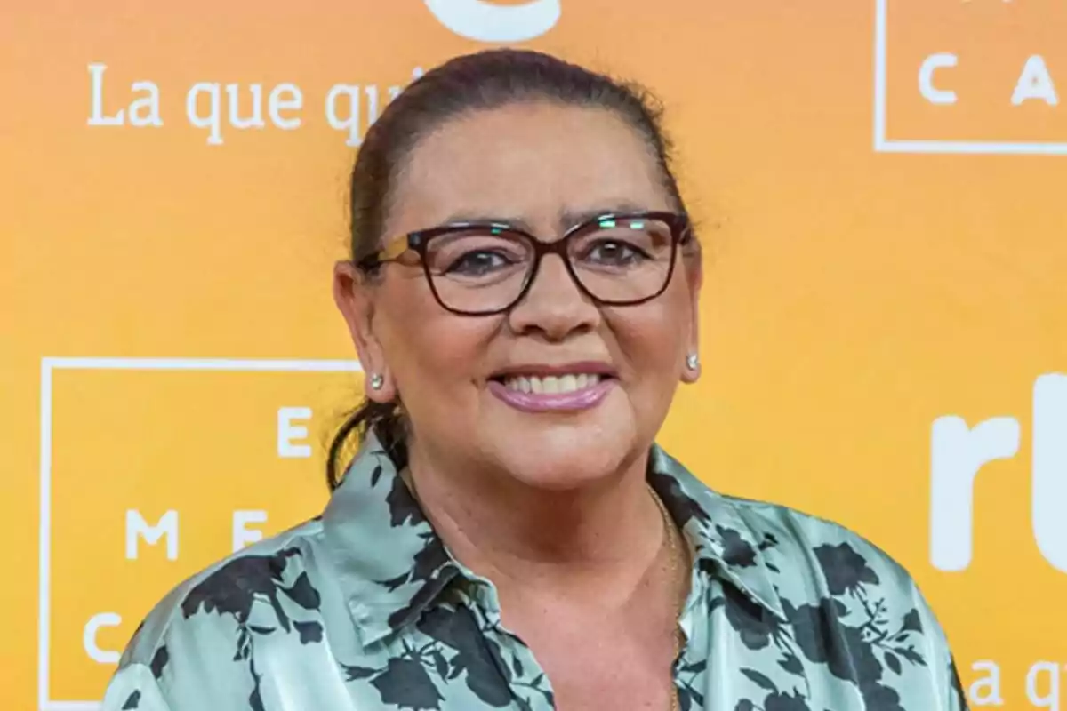 María del Monte con gafas y camisa estampada sonríe frente a un fondo naranja con texto en un photocall de RTVE.