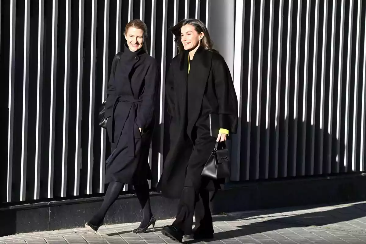 María Dolores Ocaña y la reina Letizia caminando juntas, vestidas con abrigos oscuros, frente a una pared con barras verticales. María Dolores Ocaña y la reina Letizia caminando juntas, vestidas con abrigos oscuros, frente a una pared con barras verticales.