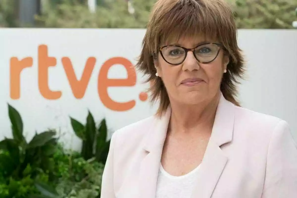 Fotografía de María Escario con gafas y chaqueta clara frente a un cartel de RTVE.