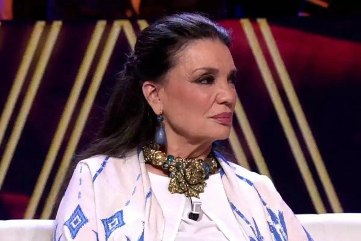 María José Cantudo con cabello oscuro y recogido lleva un collar dorado y azul y un pendiente azul, vestida con una prenda blanca con detalles azules, sentada en un entorno iluminado en De Viernes. María José Cantudo con cabello oscuro y recogido lleva un collar dorado y azul y un pendiente azul, vestida con una prenda blanca con detalles azules, sentada en un entorno iluminado en De Viernes.