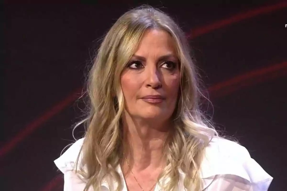 María José Galera con cabello rubio y ondulado con expresión seria, vestida con una blusa blanca, en un fondo oscuro con luces rojas en el plató de Gran Hermano.