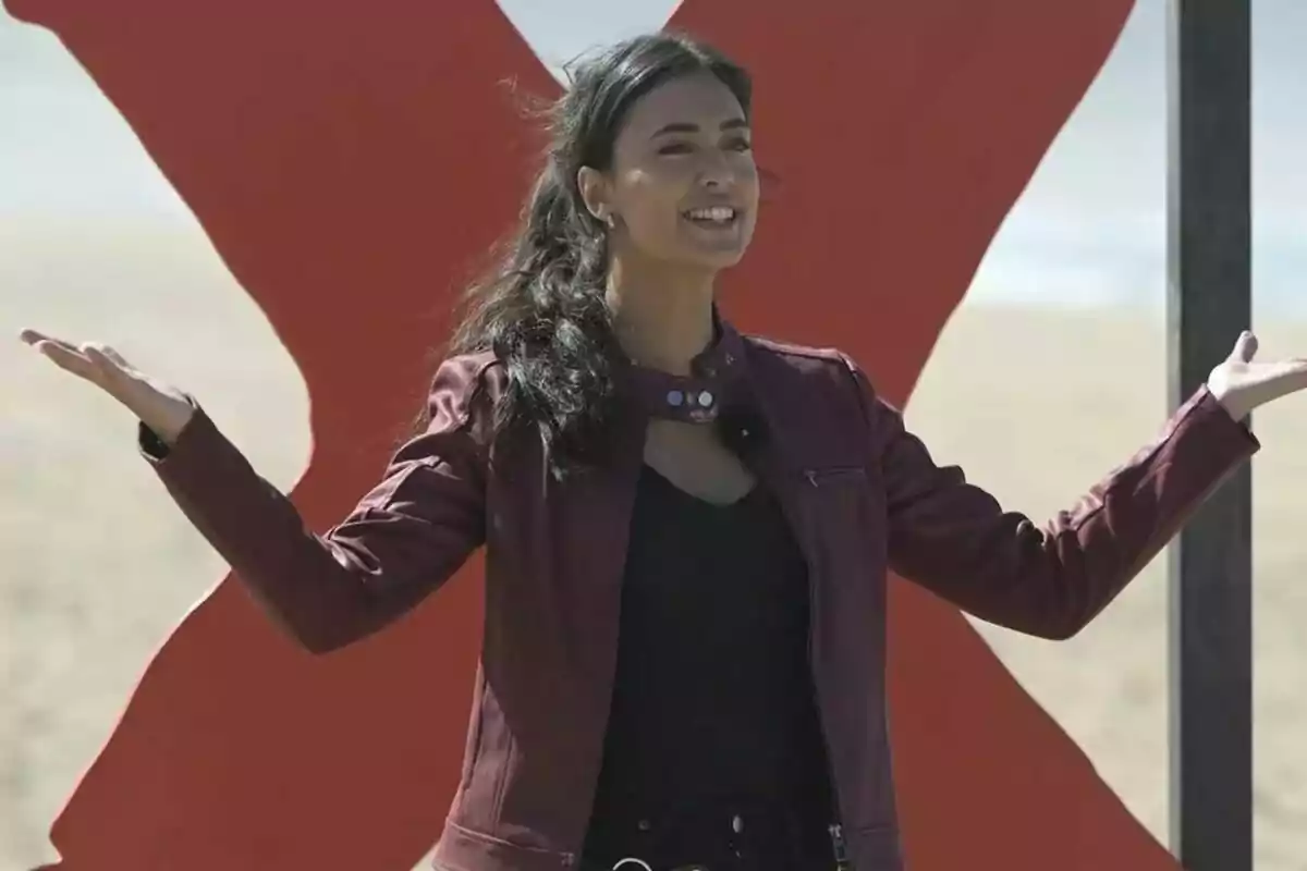 María Lamela sonriente con chaqueta roja y los brazos extendidos frente a una gran X roja en la playa.