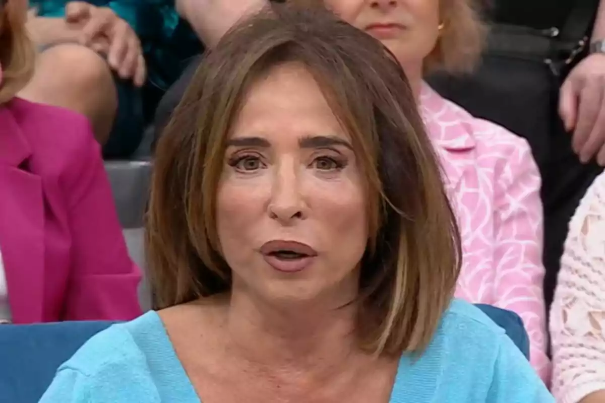 María Patiño con cabello castaño y expresión seria está sentada en un entorno de estudio de televisión en La Familia de la Tele, rodeada de otras personas.
