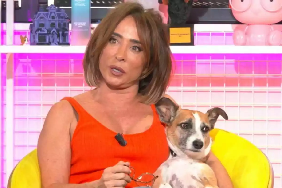 María Patiño con un vestido naranja sostiene a un perro pequeño mientras está sentada en un sillón amarillo, con un fondo de estantería decorada, en Ni que fuéramos.