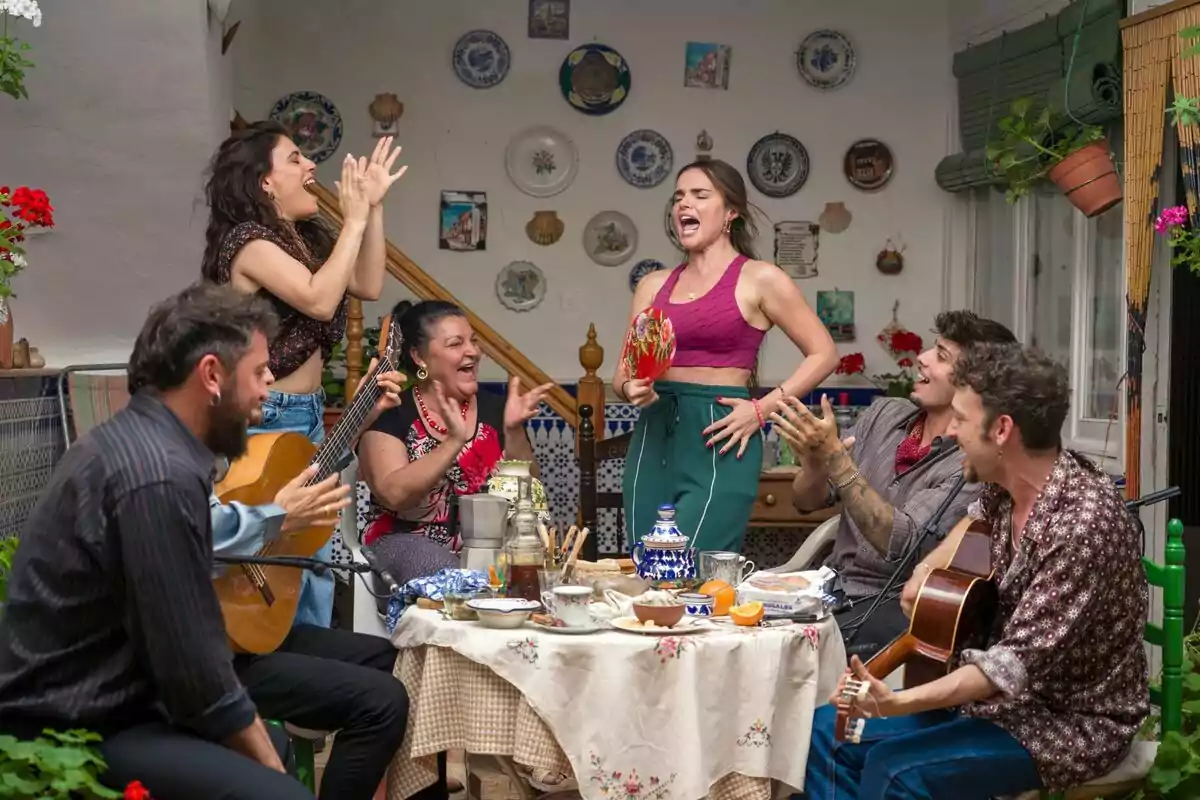 Un grupo de personas con Melody como protagonista disfruta de una reunión animada en un patio decorado con platos en la pared, mientras tocan la guitarra, cantan y aplauden alrededor de una mesa con comida y bebida en Mariliendre. Un grupo de personas con Melody como protagonista disfruta de una reunión animada en un patio decorado con platos en la pared, mientras tocan la guitarra, cantan y aplauden alrededor de una mesa con comida y bebida en Mariliendre.