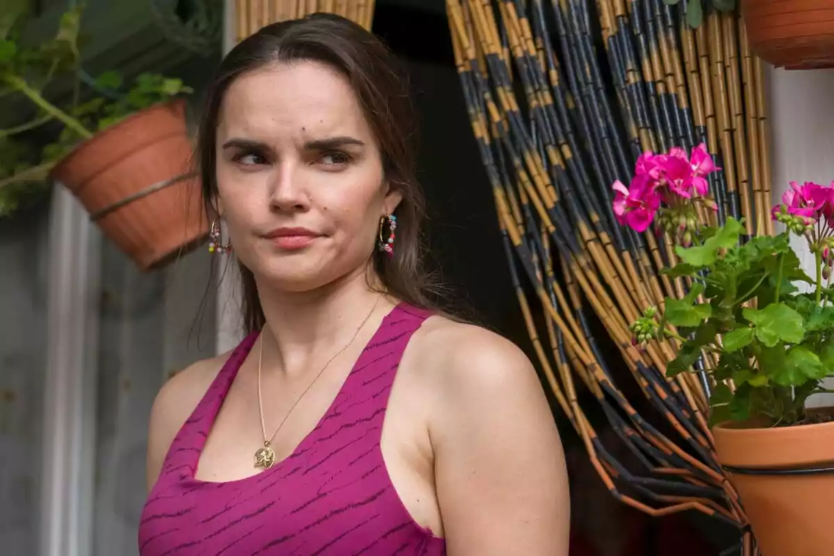 Melody con expresión pensativa usando un vestido morado y pendientes coloridos, de pie junto a una cortina de bambú y macetas con flores rosas en Mariliendre