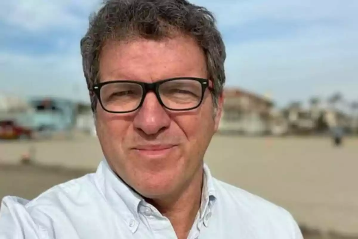 Un hombre con gafas y camisa blanca sonriendo al aire libre con un fondo borroso de edificios y cielo azul. Un hombre con gafas y camisa blanca sonriendo al aire libre con un fondo borroso de edificios y cielo azul.
