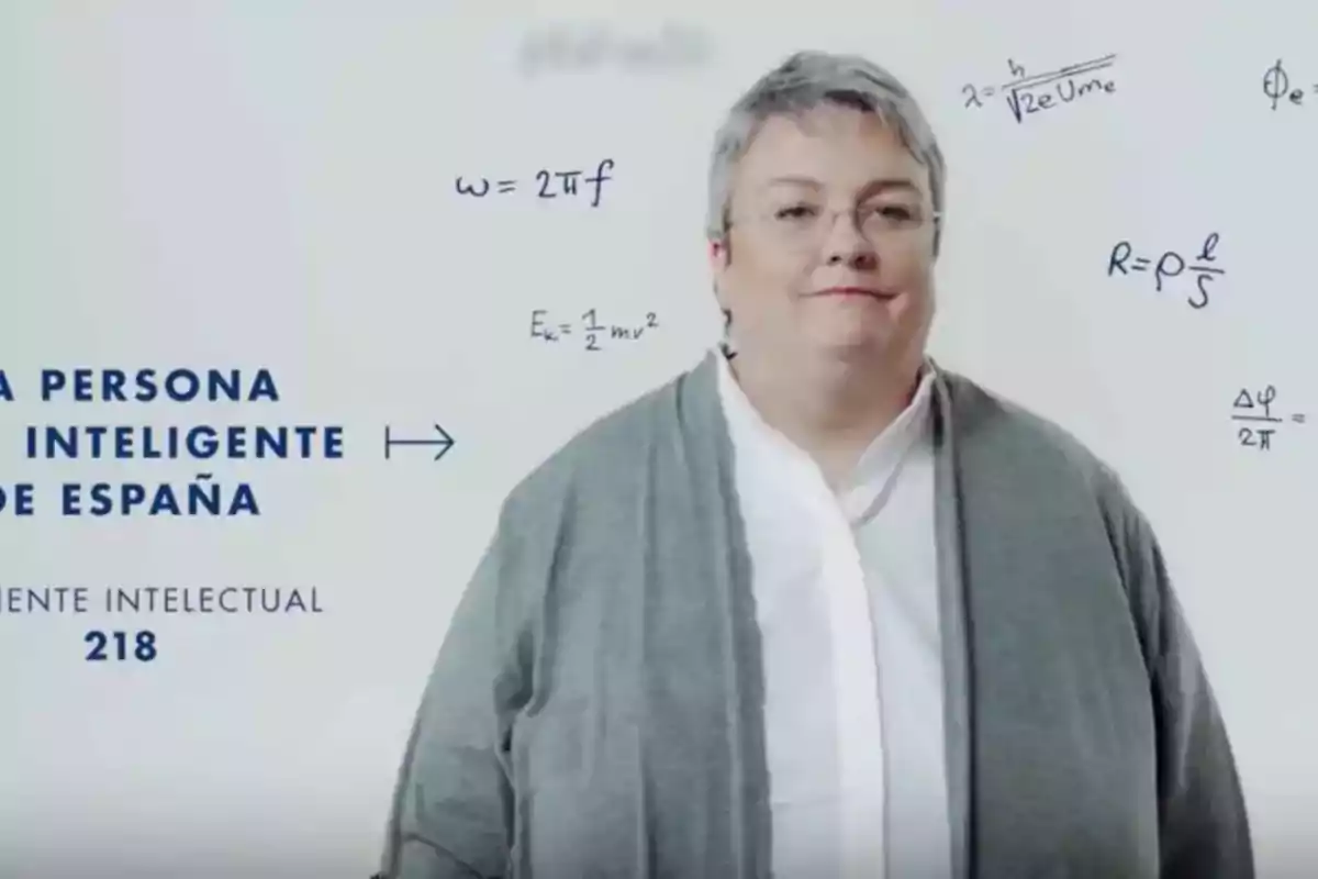 Una persona con cabello corto y canoso lleva una chaqueta gris y una camisa blanca, rodeada de fórmulas matemáticas en un fondo blanco.