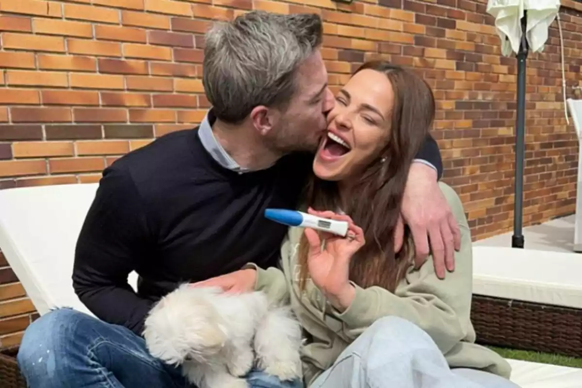 Marta Peñate y Tony Spina sonriente se abraza mientras la mujer sostiene una prueba de embarazo positiva y un pequeño perro blanco está en su regazo. Marta Peñate y Tony Spina sonriente se abraza mientras la mujer sostiene una prueba de embarazo positiva y un pequeño perro blanco está en su regazo.