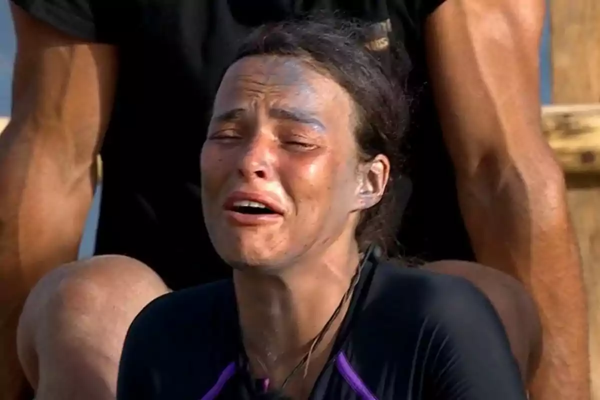 Captura de Marta Peñate con el rostro sucio y expresión de llanto, con los ojos cerrados y la boca abierta, mientras una persona en el fondo la observa en Supervivientes