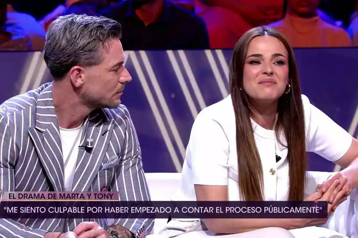 Tony Spina con chaqueta de rayas y Marta Peñate de blanco están sentados en un plató de televisión de De Viernes con público al fondo y un rótulo en pantalla que menciona el drama de Marta y Tony Tony Spina con chaqueta de rayas y Marta Peñate de blanco están sentados en un plató de televisión de De Viernes con público al fondo y un rótulo en pantalla que menciona el drama de Marta y Tony