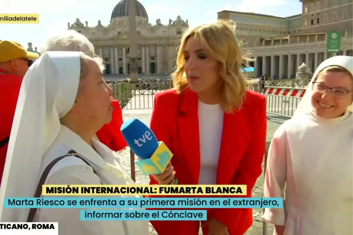 Marta Riesco con un micrófono de TVE en La Familia de la Tele entrevista a una monja en la Plaza de San Pedro en el Vaticano mientras otra monja sonríe al lado. Marta Riesco con un micrófono de TVE en La Familia de la Tele entrevista a una monja en la Plaza de San Pedro en el Vaticano mientras otra monja sonríe al lado.