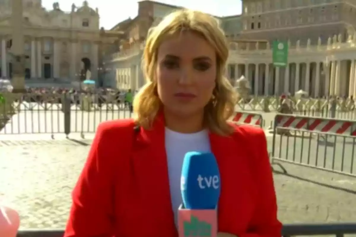 Marta Riesco con chaqueta roja sostiene un micrófono de TVE en La Familia de la Tele frente a un edificio histórico con columnas y una plaza concurrida.