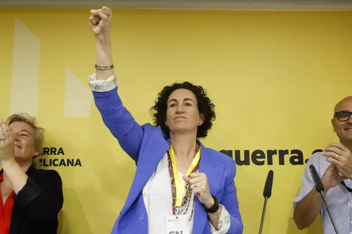 Marta Rovira de ERC levanta el puño en un evento con un fondo amarillo que tiene texto parcialmente visible.