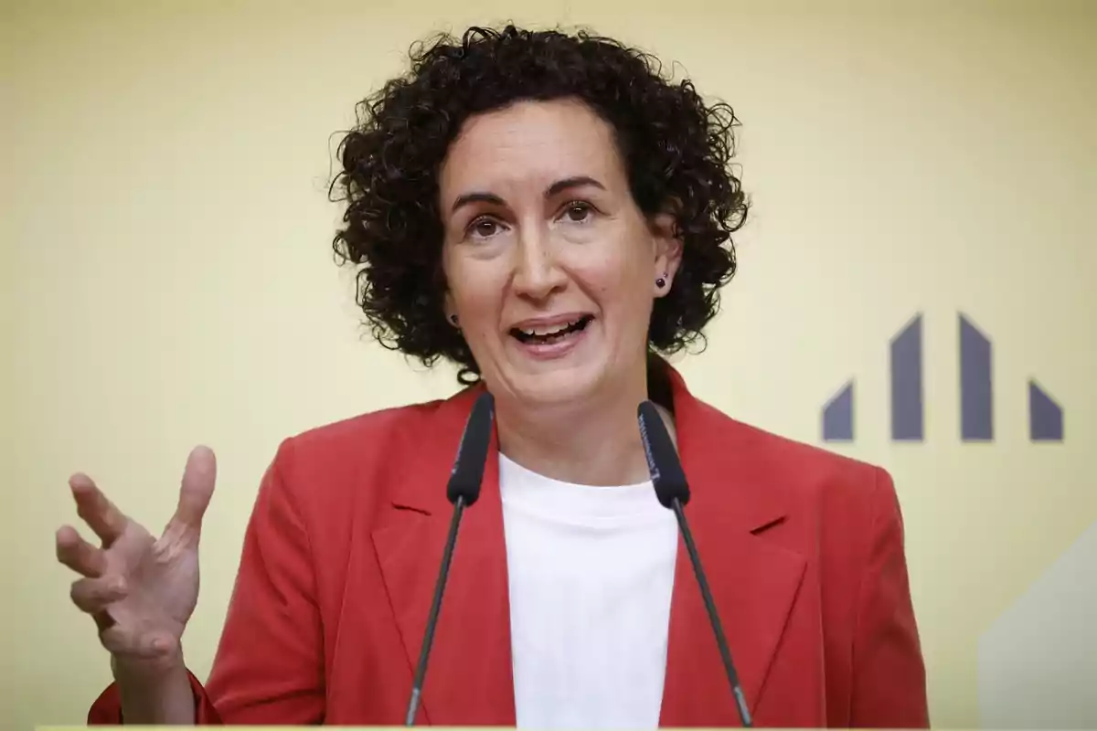 Una mujer con cabello rizado y corto, vestida con un saco rojo y una camiseta blanca, hablando frente a dos micrófonos.