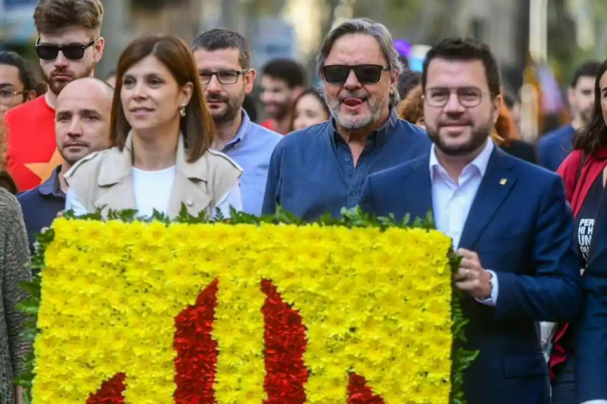 Un grupo de personas participa en una manifestación, llevando un arreglo floral con los colores amarillo y rojo.