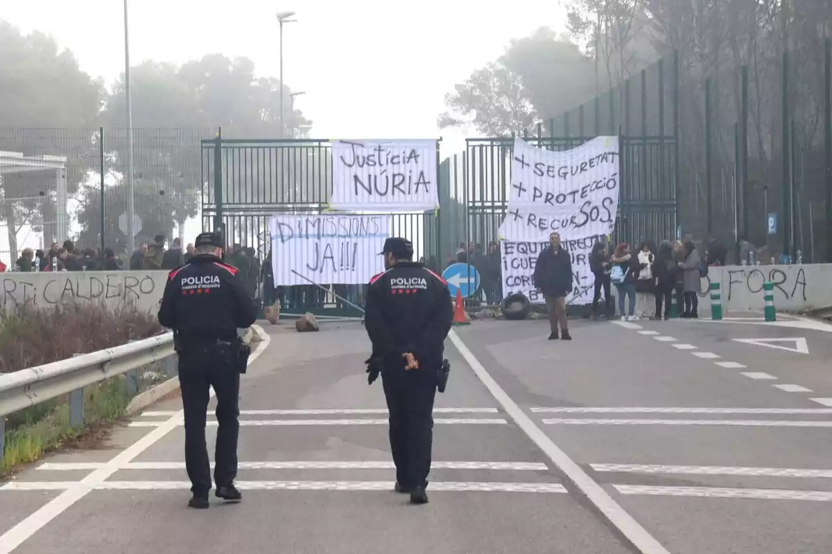 Dos Mossos d'Esquadra de espaldas delante de decenas de trabajadores en las puertas de la cárcel de Mas d'Enric con pancartas pidiendo justícia para Núria, dimisiones y más medidas de seguridad