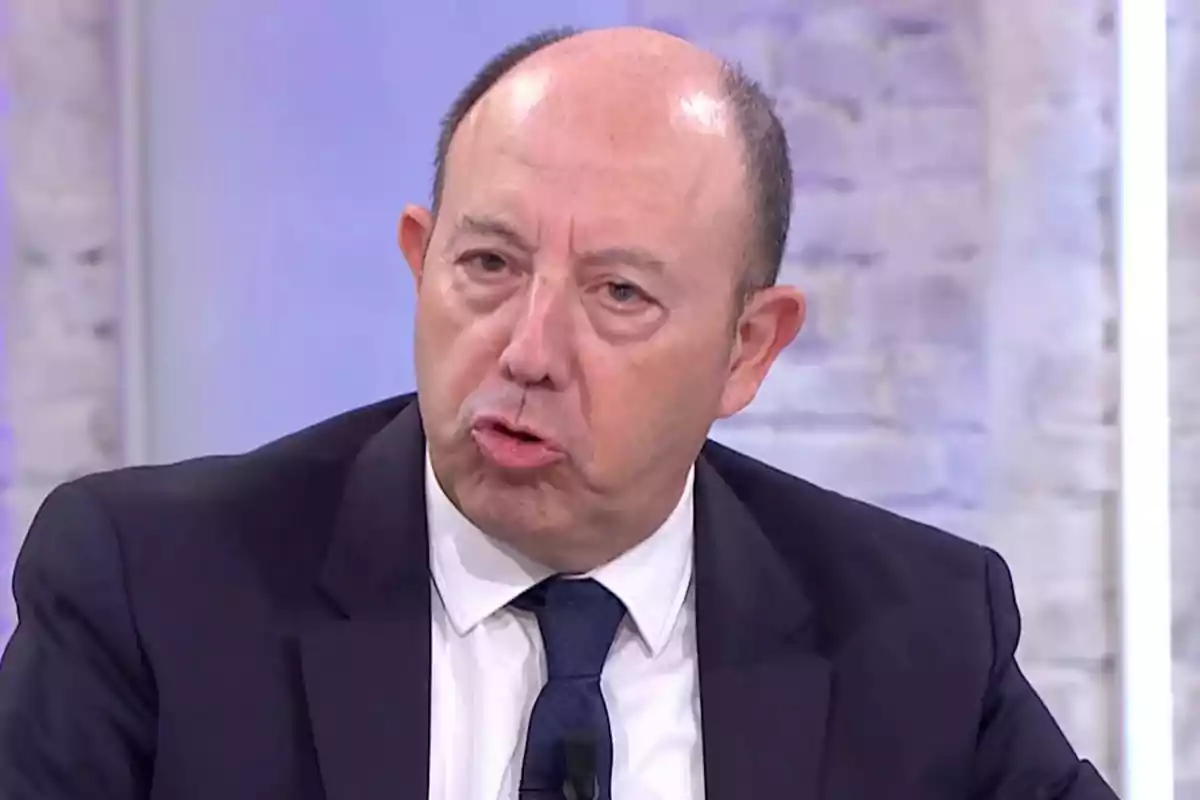 Hombre calvo de mediana edad con traje oscuro y corbata azul hablando en un estudio de televisión con fondo claro Hombre calvo de mediana edad con traje oscuro y corbata azul hablando en un estudio de televisión con fondo claro