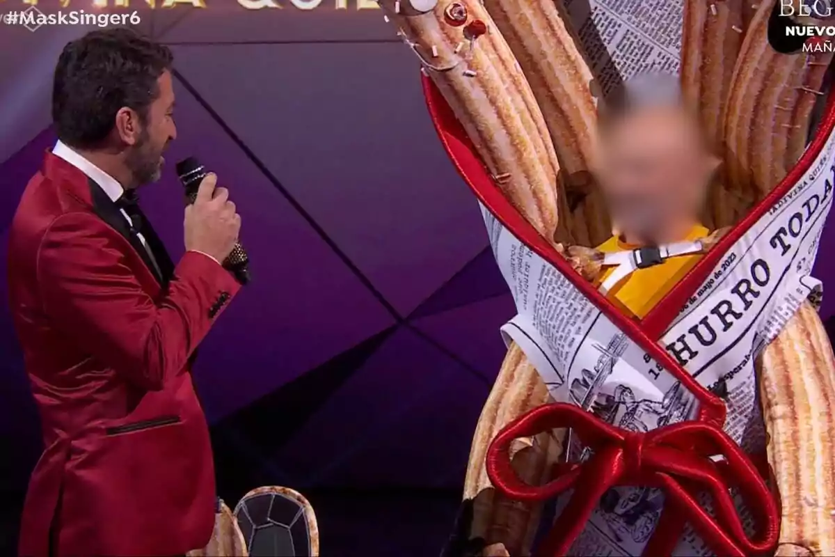 Arturo Valls con un traje rojo habla con una persona disfrazada de churro envuelta en papel de periódico en Mask Singer.