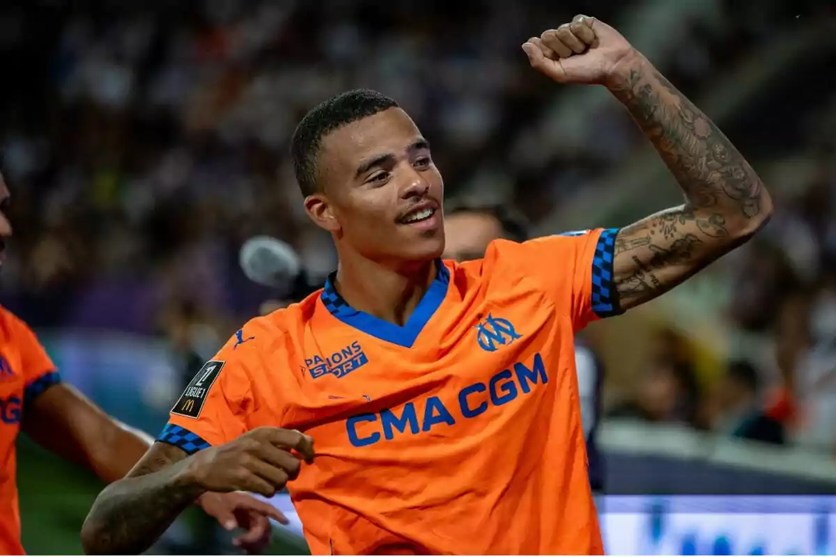 Jugador de fútbol celebrando con el puño en alto mientras viste un uniforme naranja del equipo CMA CGM. Jugador de fútbol celebrando con el puño en alto mientras viste un uniforme naranja del equipo CMA CGM.