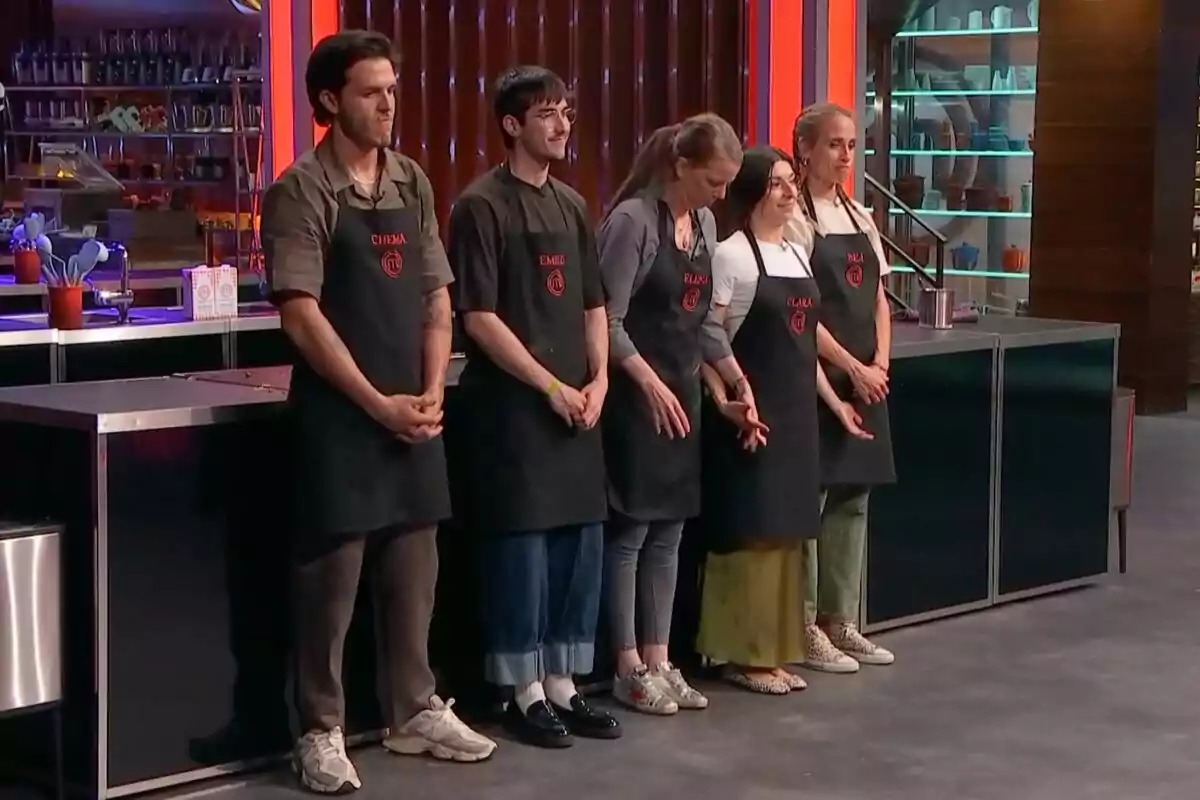 Cinco concursantes de 'Masterchef' están de pie en fila en una cocina profesional, todos llevan delantales negros con sus nombres y el logo del programa.