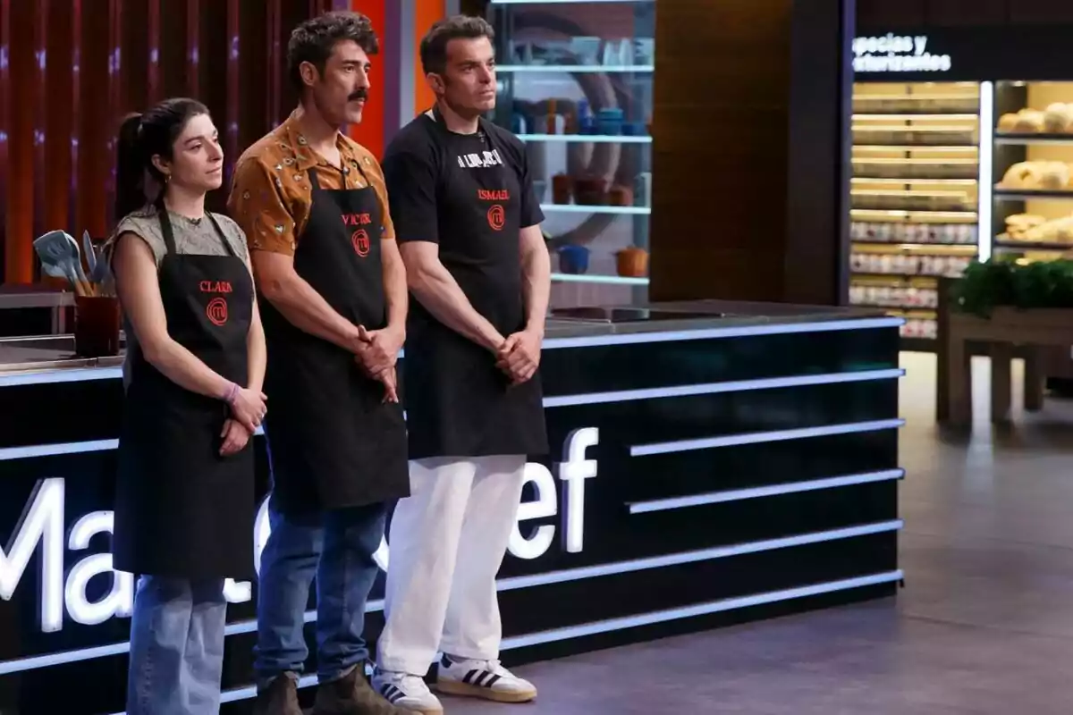 Tres personas con delantales negros están de pie en un set de cocina, participando en 'Masterchef'.