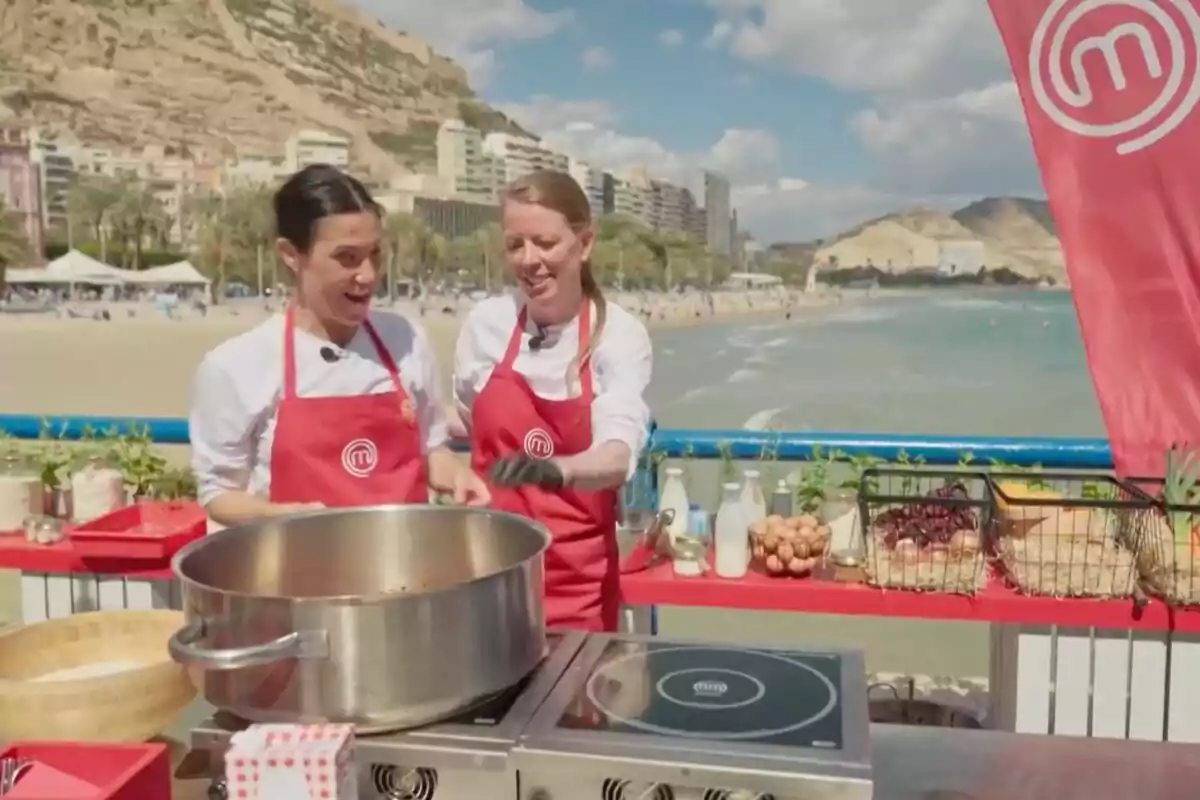 Dos personas con delantales rojos cocinan al aire libre junto a la playa, con ingredientes y utensilios sobre una mesa y edificios y montañas al fondo en 'MasterChef' Dos personas con delantales rojos cocinan al aire libre junto a la playa, con ingredientes y utensilios sobre una mesa y edificios y montañas al fondo en 'MasterChef'
