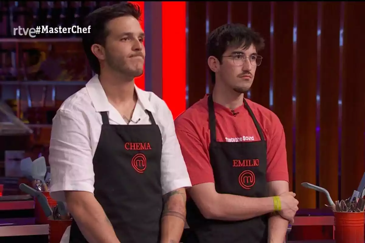 Dos concursantes de 'MasterChef' con delantales negros y nombres bordados en rojo están de pie en la cocina del programa con expresión seria.