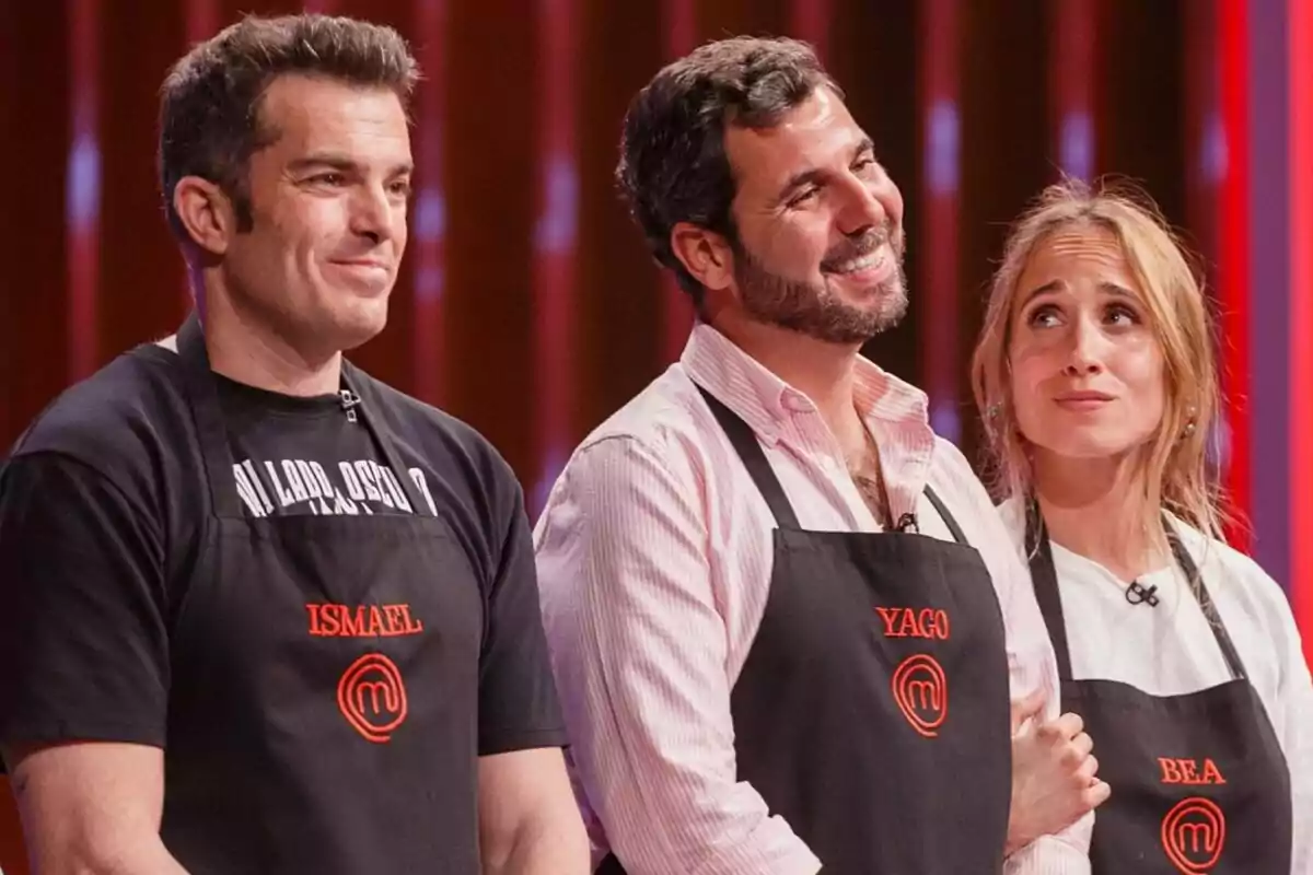 Tres concursantes de MasterChef con delantales negros y nombres bordados, posan juntos en el set del programa con expresiones de expectativa y emoción.