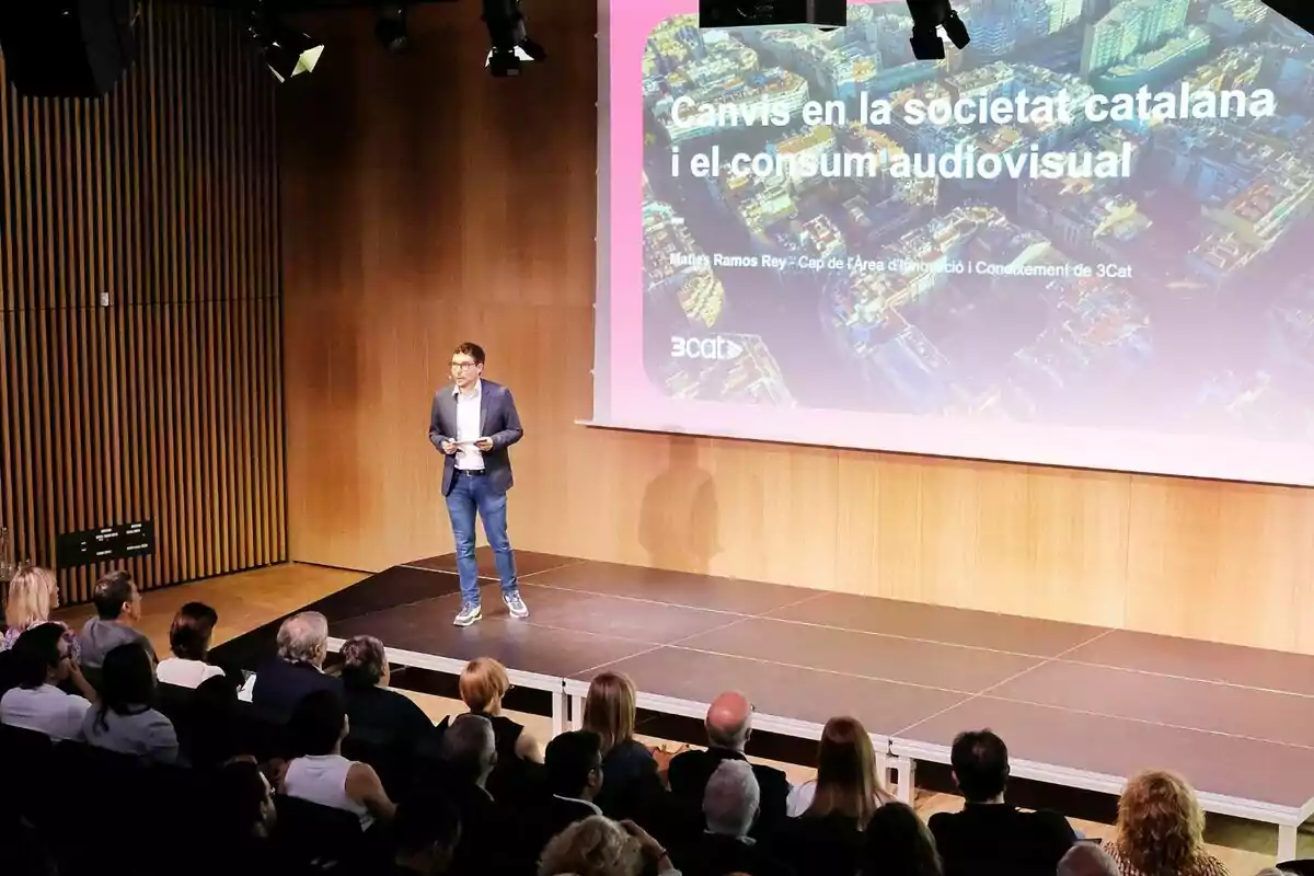 Maties Ramos de pie en un escenario da una presentación frente a una audiencia, con una pantalla grande al fondo que muestra un título sobre cambios en la sociedad catalana y el consumo audiovisual de 3Cat. Maties Ramos de pie en un escenario da una presentación frente a una audiencia, con una pantalla grande al fondo que muestra un título sobre cambios en la sociedad catalana y el consumo audiovisual de 3Cat.