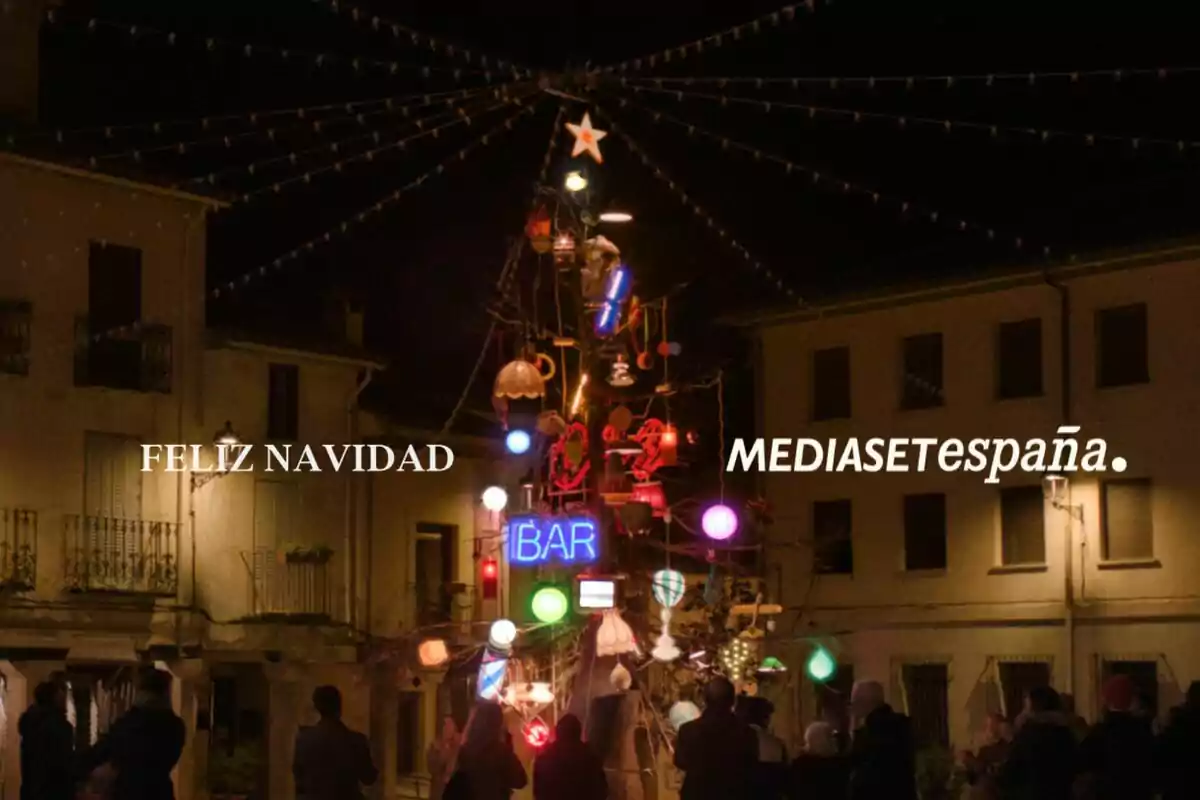 Una plaza iluminada con un árbol de Navidad decorado con luces y adornos coloridos, rodeado de personas, con las palabras "Feliz Navidad" y "Mediaset España" en el cielo nocturno.