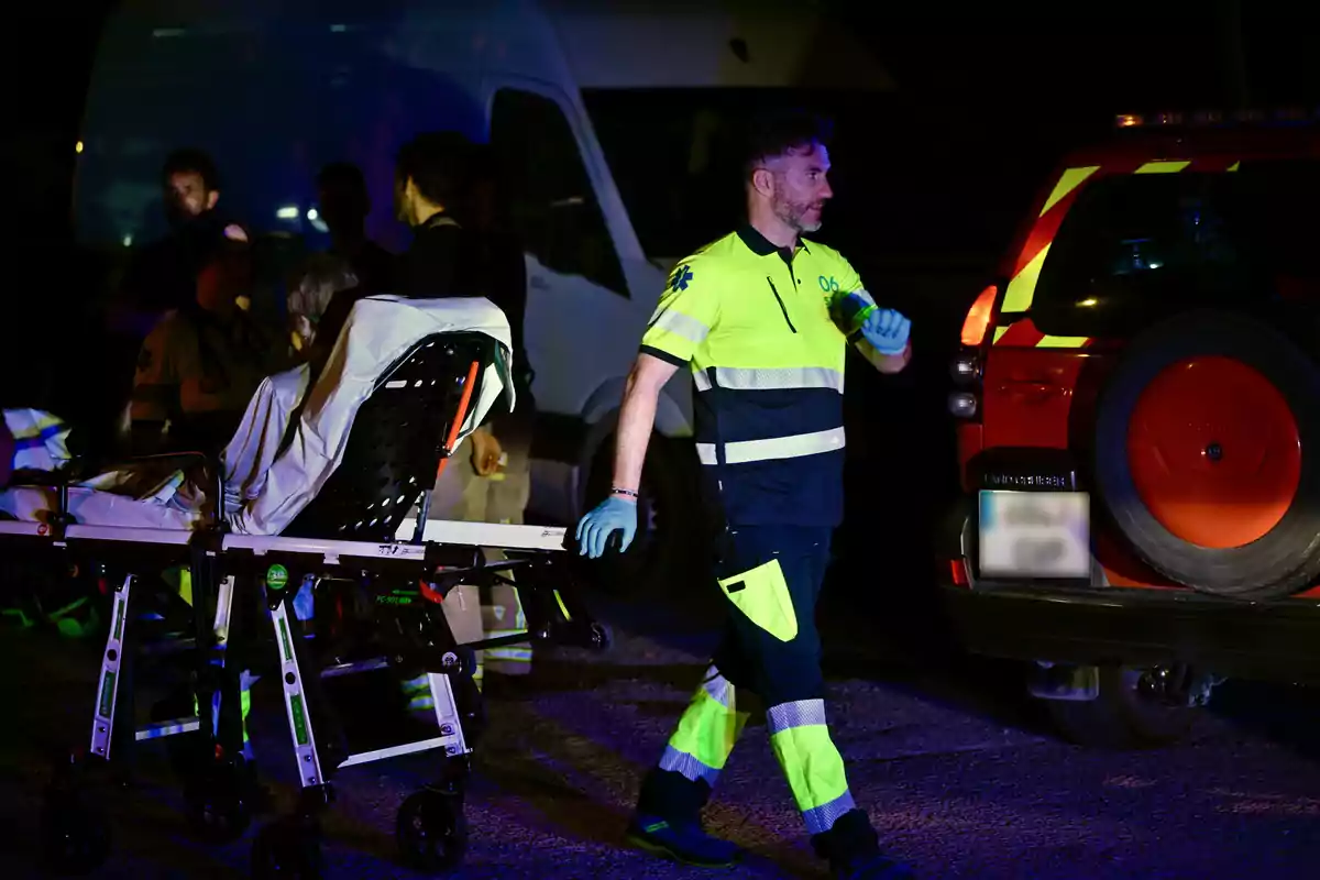 Un paramédico con uniforme reflectante empuja una camilla con una persona sentada mientras varias personas observan cerca de una ambulancia y un vehículo de emergencia durante la noche Un paramédico con uniforme reflectante empuja una camilla con una persona sentada mientras varias personas observan cerca de una ambulancia y un vehículo de emergencia durante la noche