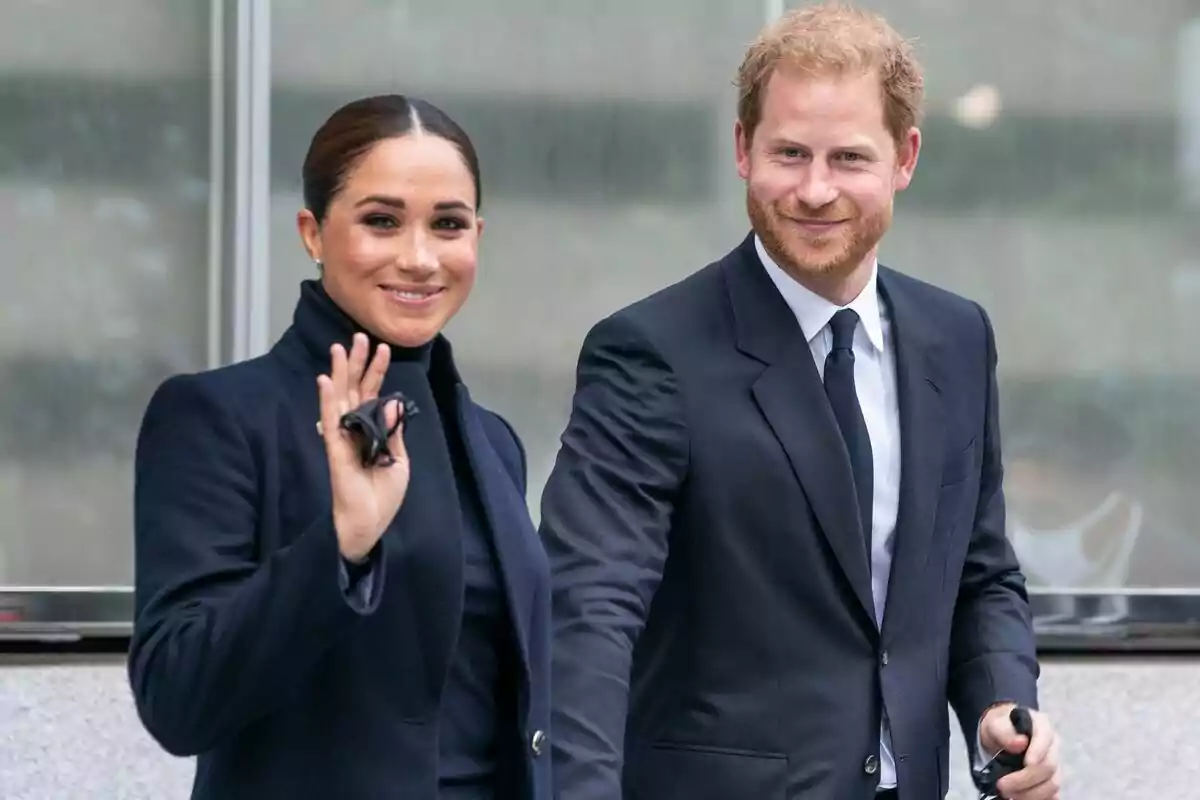 Meghan Markle y el príncipe Harry vestidos de manera formal, ambos sonriendo y caminando juntos, ella está saludando con la mano.