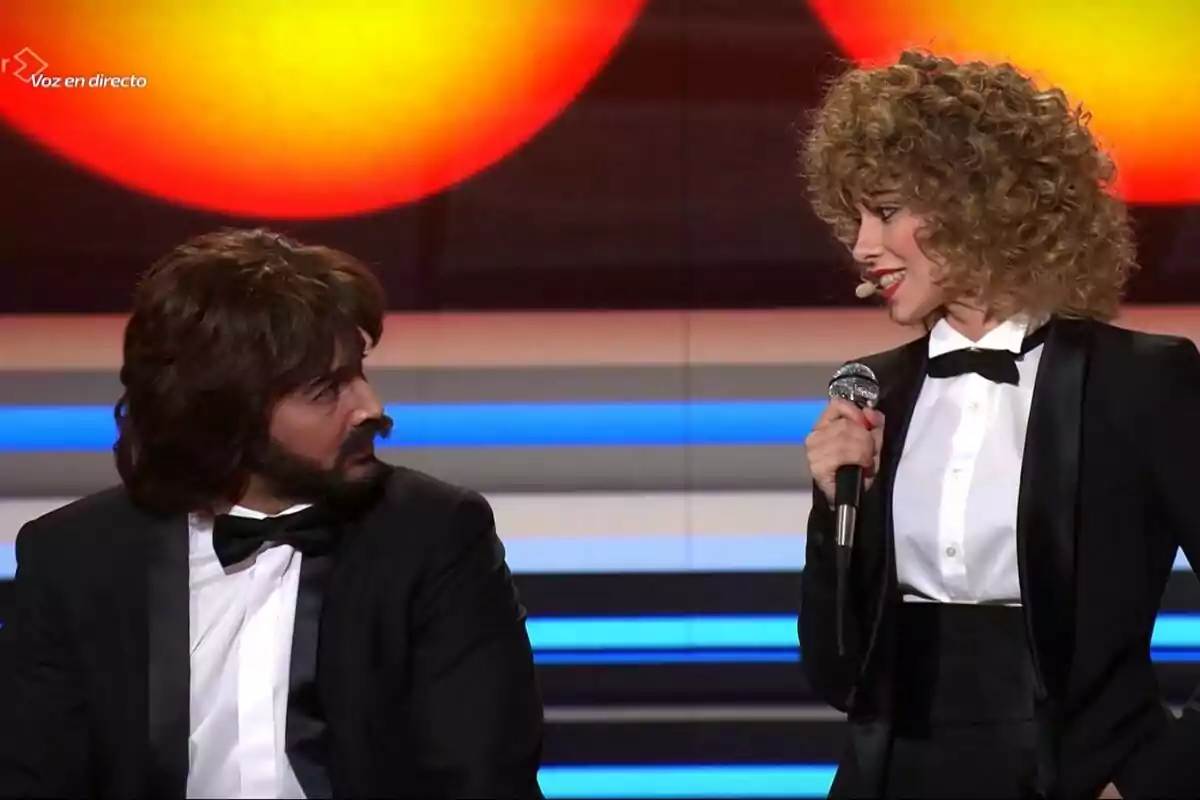 Jorge González y Melani vestidas de gala están en un escenario de Tu Cara Me Suena, una de ellas sostiene un micrófono mientras la otra la observa. Jorge González y Melani vestidas de gala están en un escenario de Tu Cara Me Suena, una de ellas sostiene un micrófono mientras la otra la observa.