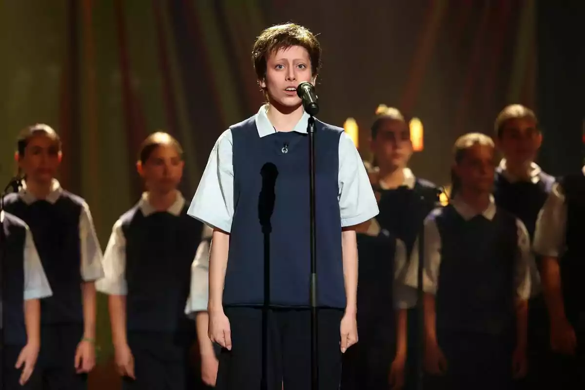 Melani vestida con uniforme escolar canta frente a un micrófono mientras un grupo de estudiantes la acompaña en el escenario iluminado en Tu Cara Me Suena.