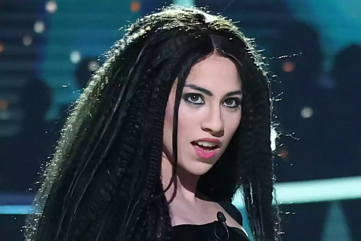 Melani con cabello largo y rizado, maquillaje llamativo y expresión intensa en un escenario iluminado en Tu Cara Me Suena.