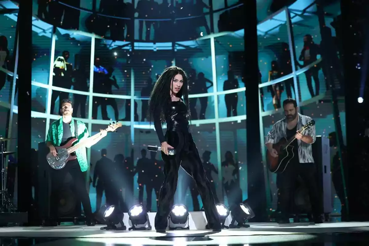 Melani García con vestuario negro y cabello rizado actuando en el escenario junto a dos músicos con guitarras y un fondo iluminado con siluetas de personas en Tu Cara Me Suena Melani García con vestuario negro y cabello rizado actuando en el escenario junto a dos músicos con guitarras y un fondo iluminado con siluetas de personas en Tu Cara Me Suena