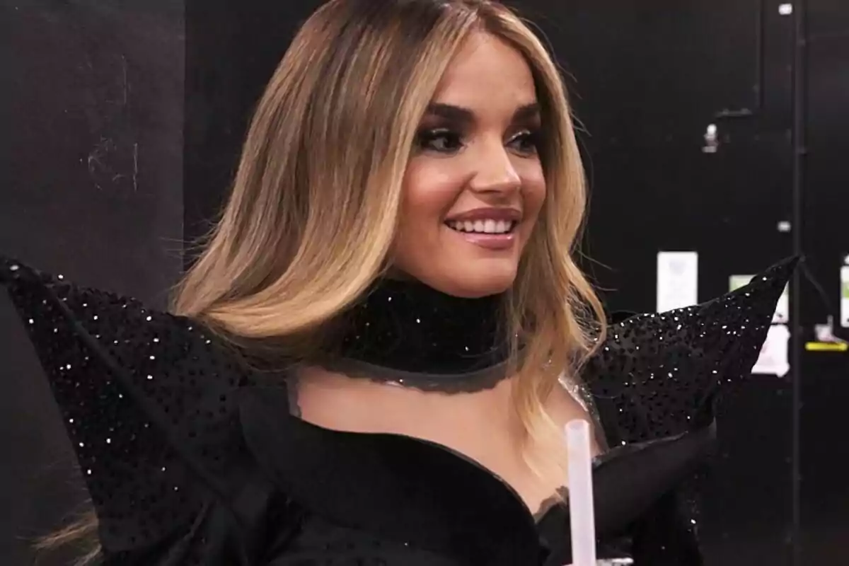 Melody con cabello largo y rubio sonriendo, vistiendo un traje negro con detalles brillantes y hombreras puntiagudas.