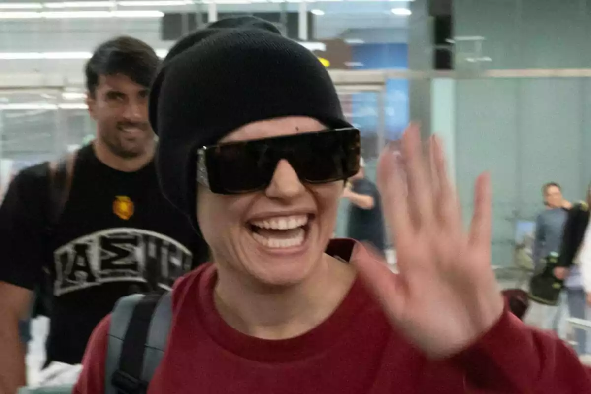 Melody sonriente con gafas de sol y gorro negro saluda mientras camina en su llegada al aeropuerto.