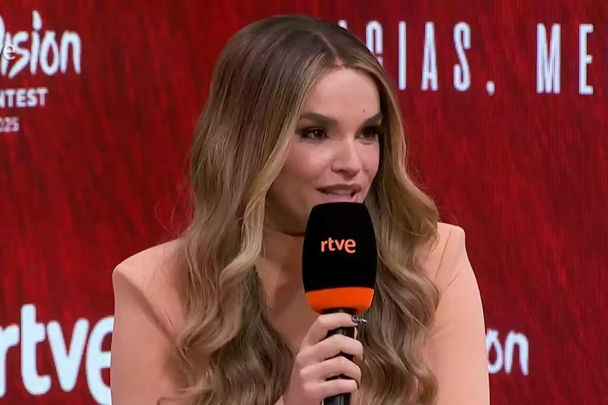 Melody de cabello largo y ondulado hablando frente a un micrófono de rtve con fondo rojo y letras blancas en Eurovisión. Melody de cabello largo y ondulado hablando frente a un micrófono de rtve con fondo rojo y letras blancas en Eurovisión.