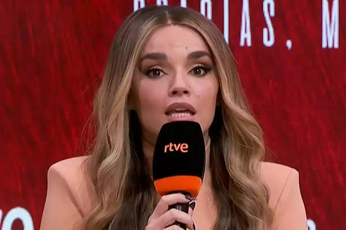 Melody con cabello largo y ondulado sostiene un micrófono negro con el logo de rtve mientras habla frente a un fondo rojo durante la rueda de prensa de Eurovisión.
