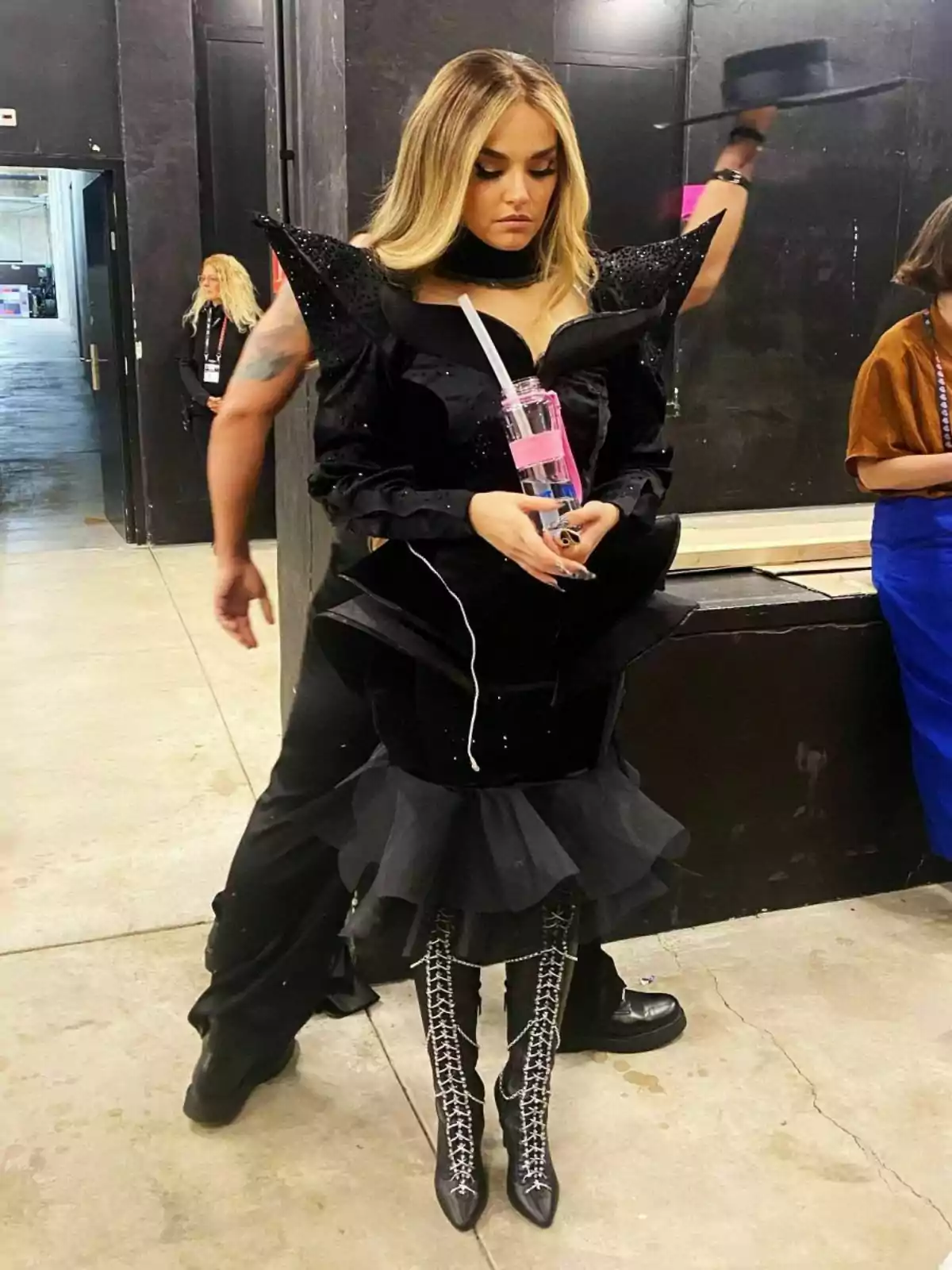 Melody con un vestido negro de diseño único y botas altas con cordones sostiene una botella de agua mientras está de pie en un entorno de backstage. Melody con un vestido negro de diseño único y botas altas con cordones sostiene una botella de agua mientras está de pie en un entorno de backstage.